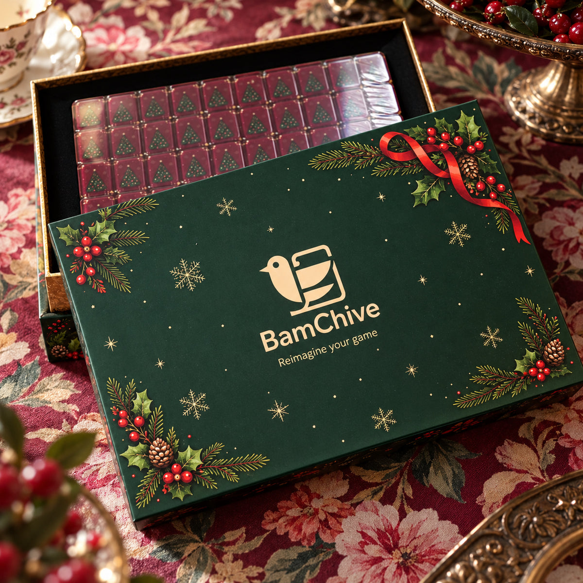 Green BamChive Christmas mahjong gift box with gold branding, holly, and red ribbons, with tree-backed tiles visible behind the lid.